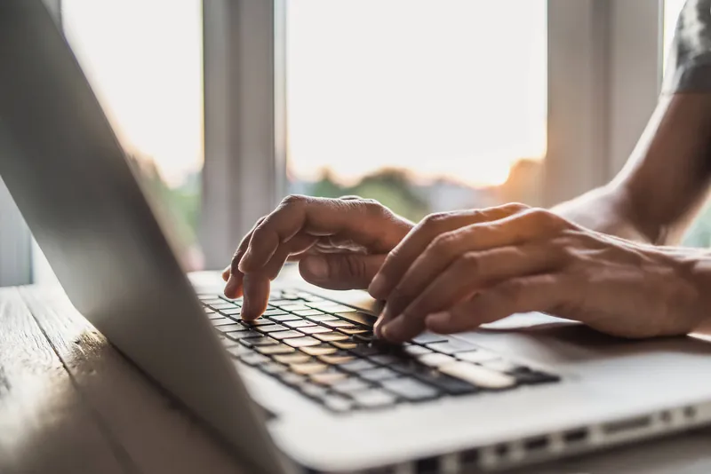 Hands typing on a laptop at a sunlit desk